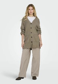 Cardigan en tricot beige avec un col en V, des manches longues et une fermeture à boutons. Il dispose de deux poches à l'avant et d'un tissu doux et texturé.