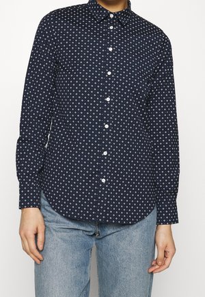 Person wearing a navy button-up shirt with white geometric patterns and light blue jeans, standing against a plain white background.