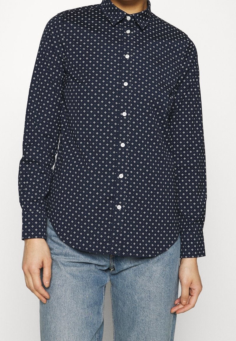 Personne portant une chemise bleu marine à boutons avec des motifs géométriques blancs et un jean bleu clair, debout devant un fond blanc uni.