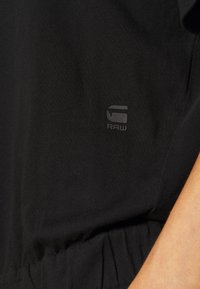 Detail černé látky s malým šedým logem G-Star RAW poblíž bočního švu a s částečně viditelnou rukou na pravém okraji.