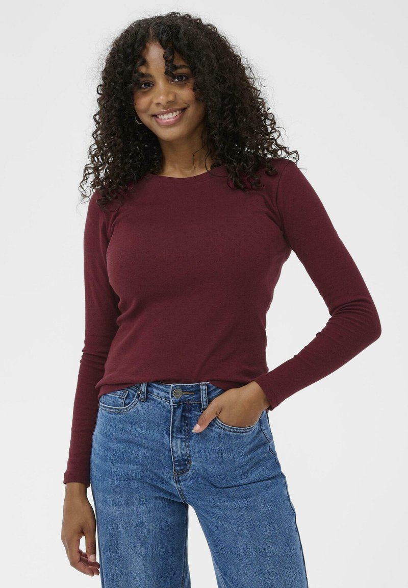 Maroon long-sleeve top with a round neckline, made of smooth fabric. Paired with high-waisted blue denim jeans. Model's hand resting on hip.