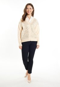 Bežový cardigan s výšivkami z flitrů, zapínání na knoflíky a žebrovanými rukávy, spárovaný s černými slim-fit kalhotami a nahými vysokými podpatky.