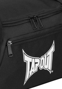 Bolsa de deporte de tela negra con un bolsillo frontal con cremallera, cremalleras de metal y un prominente diseño del logo blanco "TAPOUT". Construcción duradera y práctica.