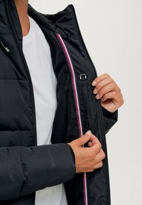 Zwarte gewatteerde jas met rits en roze accentstrepen. Glanzende textuur en verstelbare capuchon, met handen die de rits bedienen.