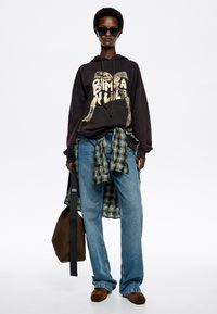Sudadera negra con estampado gráfico dorado, vaqueros azules anchos, camisa de cuadros verde atada a la cintura, gafas de sol negras, bolso marrón y zapatos marrones.