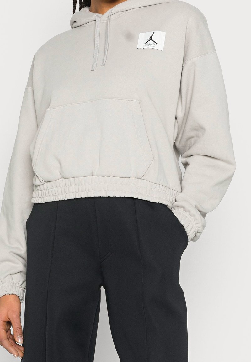 Personne portant un sweat à capuche court gris clair avec poche kangourou à l'avant et écusson logo noir, accompagné d'un pantalon foncé taille haute, une main dans la poche.