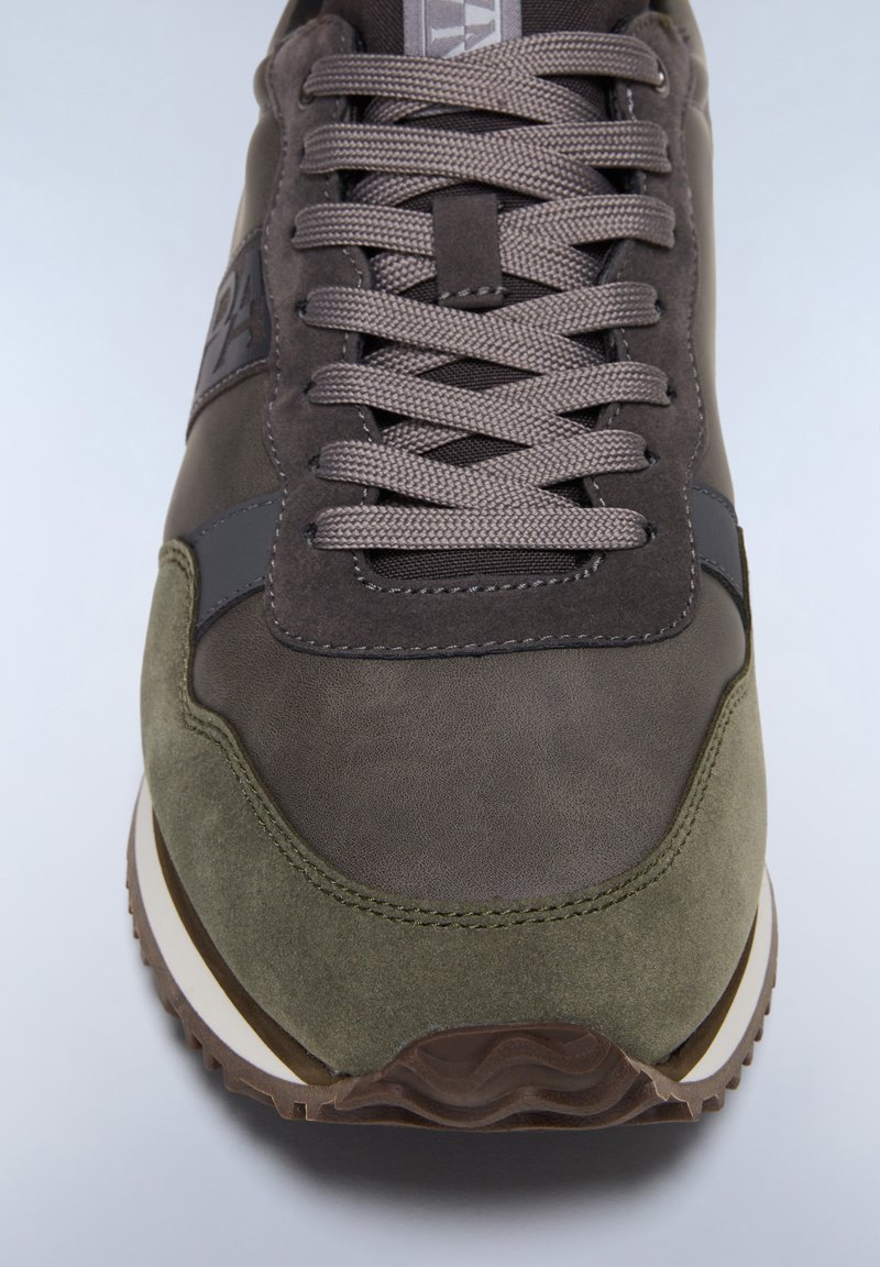 Zapatillas de color verde oliva oscuro con materiales de ante y cuero, con puntera redonda, cordones grises y una suela de goma marrón texturizada.