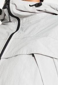 Light gray jacket featuring a textured fabric, adjustable hood, black zipper, and overlapping flap detail. Interior lining is smooth.