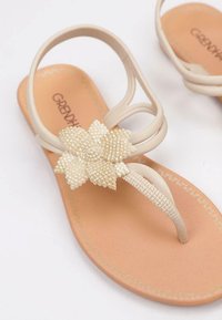 Chanclas beige con correas redondeadas, acento de flor texturizada y plantilla marrón claro con un sutil detalle dorado.