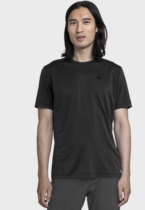 Mann mit langen schwarzen Haaren, der ein schlichtes schwarzes T-Shirt mit kurzen Ärmeln und dunkle Hose trägt, steht vor einem schlichten hellen Hintergrund.