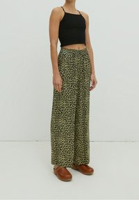 Persona che indossa pantaloni a palazzo con stampa leopardata, top corto nero e zoccoli marroni, in piedi su uno sfondo bianco semplice.
