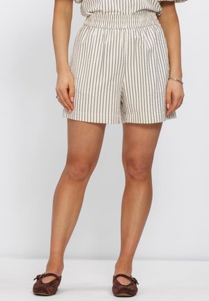 Vrouw draagt beige korte broek met verticale zwarte strepen en bruine platte schoenen, staand tegen een effen witte achtergrond.