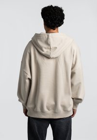 Beige oversized hoodie med en stor hætte, sænkede skuldre og ribbede manchetter. Blød tekstur og afslappet pasform, båret med mørke jeans.