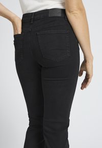 Inimene, kes kannab musti teksaseid, tagataskud on nähtavad, üks käsi on taskus ja teine ripub kõrval.