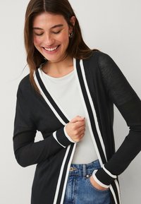 Schwarze Strickjacke mit weißen Streifen entlang des Kragens und der Bündchen, aus leichtem Stoff, offenes Design, kombiniert mit einem weißen Oberteil und Jeans.