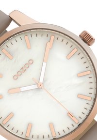 Rose gold watch with a round face, white mother-of-pearl dial, rose gold hour markers, and a beige band. Features minimalist design and a crown.