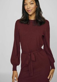 Burgundy långärmad klänning i mjukt material har en figurnära midja med ett knytbälte, räfflade ärmslut och en enkel rund halsringning.