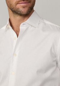 Camicia di cotone bianca con colletto a punta, caratterizzata da una sottile cucitura blu, e bottoni in plastica. Texture liscia, vestibilità su misura.