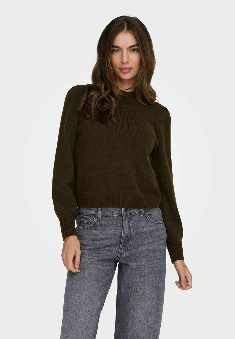 Femme aux longs cheveux bruns portant un pull vert olive foncé et un jean taille haute gris sur fond blanc.