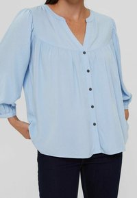 Blouse bleu clair à boutons, avec un décolleté en V, un design plissé et des manches bouffantes. Présente des boutons sombres et une coupe décontractée.