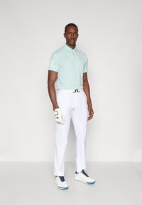 Lyseblå polo skjorte med krage og teksturerte detaljer på ermene, kombinert med hvite bukser og hvite golfsko. Holder en golfhanske.