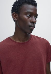 Bordeaux-farbenes T-Shirt mit kurzen Ärmeln und rundem Ausschnitt, aus weicher Stoff gefertigt, präsentiert von einer Person mit kurzen, lockigen Haaren vor einem schlichten Hintergrund.