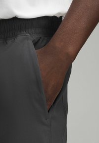 Pantaloni sportivi grigio scuro con cintura elastica e tasche laterali, mostrati con un primo piano di una mano che riposa in una tasca.
