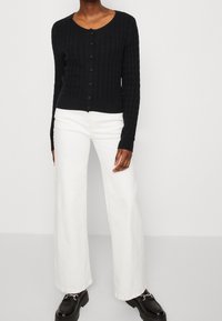 Czarny dzianinowy kardigan z fakturowanym wzorem, z okrągłym dekoltem i zapinany na guziki, zestawiony z białymi szerokimi spodniami i czarnymi butami.