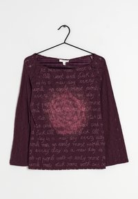 Lila spetstopp med trekvartsärmar, med ett textmönster och en central blommig design i mörkare lila nyanser.