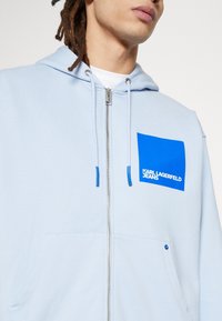 Felpa con cappuccio azzurro chiaro con cerniera e tasca frontale, con una grafica quadrata blu e testo bianco "KARL LAGERFELD JEANS." Cappuccio con coulisse.