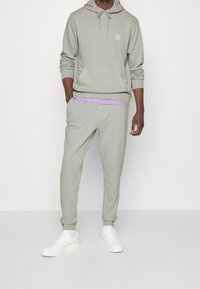 Felpa verde chiaro con tasca frontale, pantaloni jogger verdi chiari coordinati, sovrapposti a una maglietta viola, abbinati a sneaker bianche.