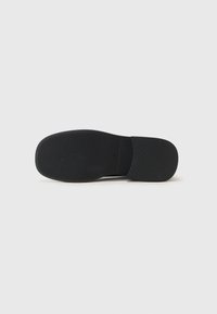 Suela de zapato negro tipo slip-on con un acabado de goma texturizada, que presenta surcos horizontales para un mejor agarre y un diseño de punta redondeada.