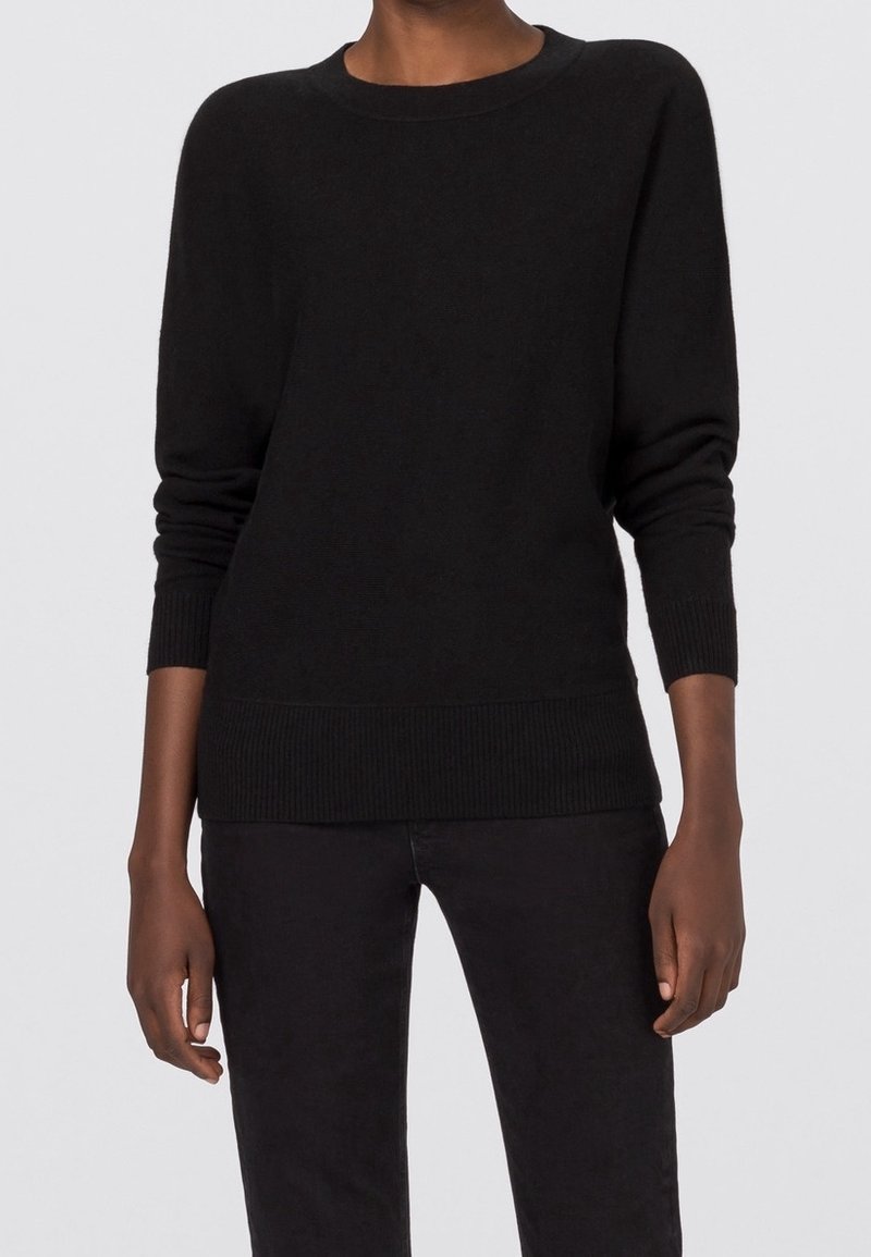 Personne portant un pull à manches longues noir uni avec des poignets et un ourlet côtelés, associé à un pantalon noir, debout devant un fond neutre.