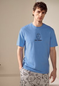 Blå bomullströja med korta ärmar, som har en svart grafik av en karaktär och texten "NOT TODAY" på bröstet. Matchad med grå mönstrade shorts.