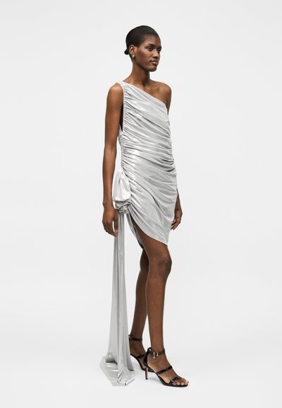 Γυναίκα με ασημένιο μίνι φόρεμα ενός ώμου με ρυτιδώσεις, μακρύ πλάι drape και μαύρα ψηλοτάκουνα σανδάλια, στέκεται σε μια απλή φόντο.