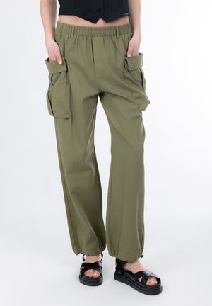 Olijfgroene cargobroek met elastische tailleband, zij- en patchzakken en taps toelopende boorden. Gecombineerd met zwarte sandalen op blote voeten.