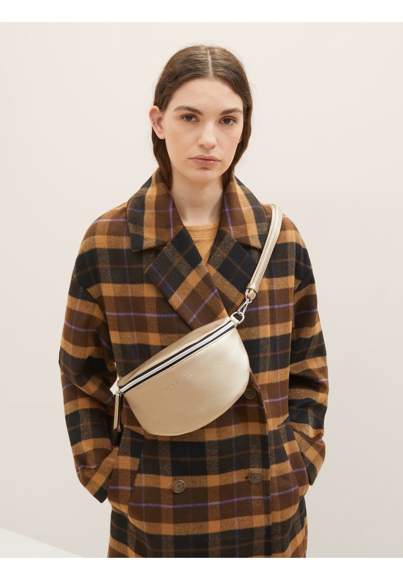 Beige læder crossbody taske med en buet form, der har en sort lynlås detalje og en subtil præget logo. Båret over en ternet brun frakke.