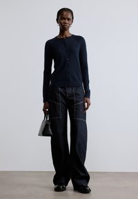 Tmavomodrý pletený sveter na gombíky v kombinácii so širokými tmavými denimovými nohavicami s kontrastnými švami. Modelka drží malú čiernu kabelku.