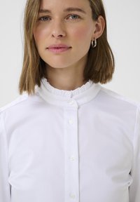 Vit skjorta med uppknäppt krage, tillverkad av slät tyg. Har genomskinliga knappar och en figursydd design som framhäver ett rent utseende.