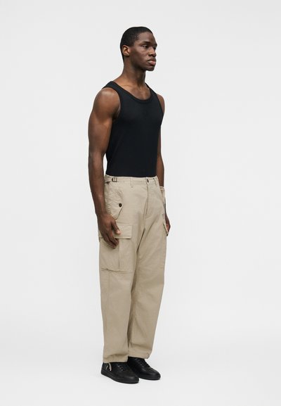 Άνδρας με μαύρο φανελάκι, μπεζ παντελόνι cargo με τσέπες και μαύρα αθλητικά παπούτσια, στέκεται μπροστά σε απλό λευκό φόντο.