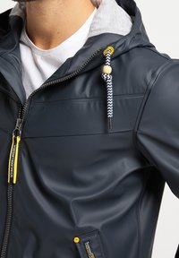 Homme portant une veste imperméable bleu marine avec doublure de capuche grise, cordon rayé avec perle en bois, et tirette de fermeture éclair et bouton avec accents jaunes.