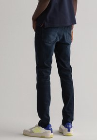 Mørkeblå jeans med slim fit, med baklommer og sømdetaljer, i kombinasjon med lyse sneakers med blå og gule aksenter.