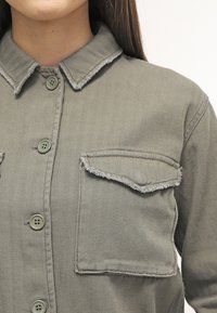 Kaki denimskjorta med fiskbensstruktur, knappstängning fram, och en bröstficka med slitna kanter. Spetsig krage och gröna knappar.