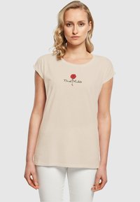 Tricou din bumbac bej, cu mâneci scurte, având un grafic cu o rose roșie și textul negru "Frida Kahlo" centrat sub design.