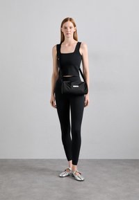 Top corto nero, leggings coordinati e una piccola borsa a tracolla nera. Scarpe argentate e uno sfondo neutro completano l'outfit.