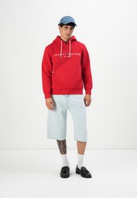 Uomo che indossa una felpa rossa Tommy Hilfiger, pantaloni corti di denim chiaro fino al ginocchio, calze bianche, mocassini neri e un cappellino da baseball blu, in piedi di fronte a uno sfondo bianco.