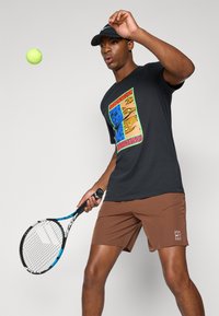Black t-shirt with colorful graphic design featuring animal and abstract patterns. Model wears brown shorts, holding a tennis racket, and a cap.