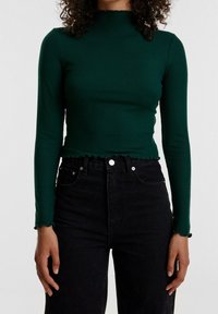 Ribbed green long-sleeve top with a high neckline and scalloped edges, paired with high-waisted black jeans featuring button accents.