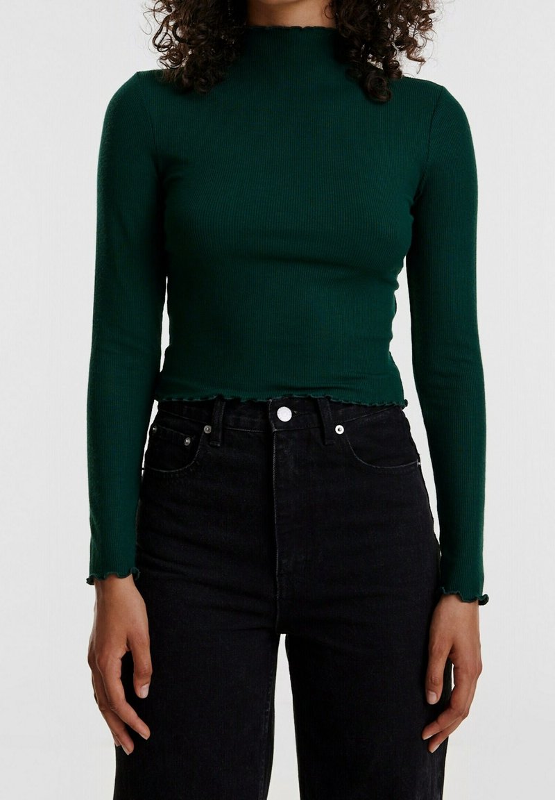 Ribbed green long-sleeve top with a high neckline and scalloped edges, paired with high-waisted black jeans featuring button accents.