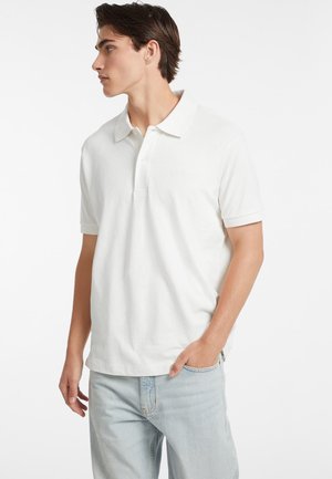 Polo bianco in cotone, con colletto classico e maniche corte. Vengono indossati pantaloni in denim chiaro, che offrono un look informale.